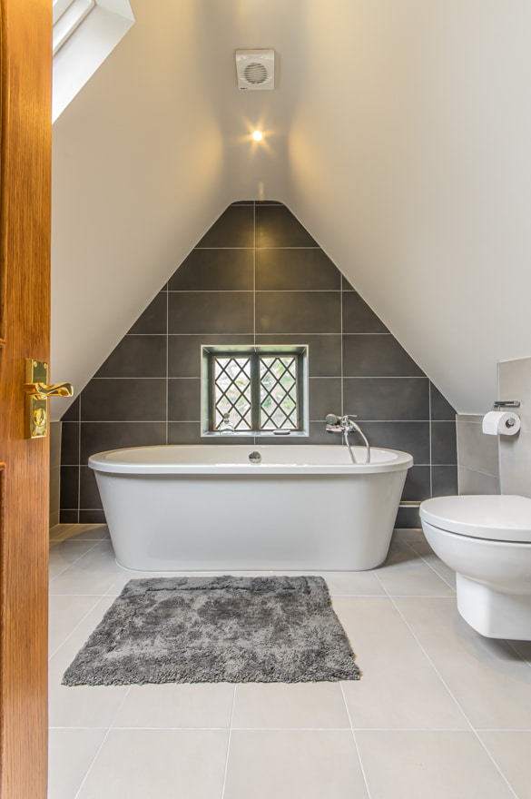 Bathroom door open showing a free standing bath against an apex wall with a window in it and wall tiles that are grey. There is a grey rug on white tiles and the celing and walls are white.